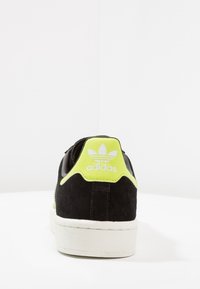 Svart mockaskor med en limegrön hälflik, vit gummisula och präglad Adidas-logotyp baktill. Ren och modern design.