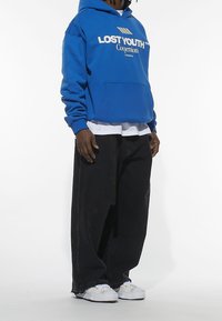 Modrá mikina s kapucí s nápisem "LOST YOUTH", kapsa vpředu, vrstvená přes bílé tričko. Nošená s volnými černými kalhotami a bílými botami.