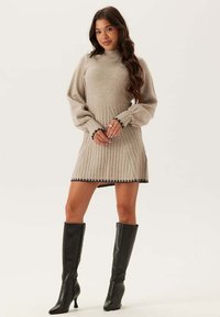 Beige ribbelgebreide jurk met een hoge halslijn, pofmouwen en zwarte afwerking aan de zoom. Gecombineerd met zwarte laarzen tot de knie.
