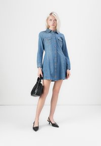 Denim shirt dress, long sleeves, buttoned front, two chest pockets, relaxed fit, light blue color, paired with a black bag and heels.