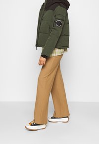 Persona che indossa una giacca a vento verde oliva, una camicia a quadri, pantaloni larghi color cammello e sneakers platform nere, bianche e gialle, in posizione laterale.