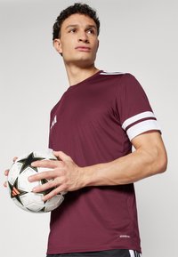 Bordeaux sportshirt met witte gestreepte mouwen, vastgehouden door een persoon met korte krullen, die ook een zwart-witte voetbal vasthoudt.