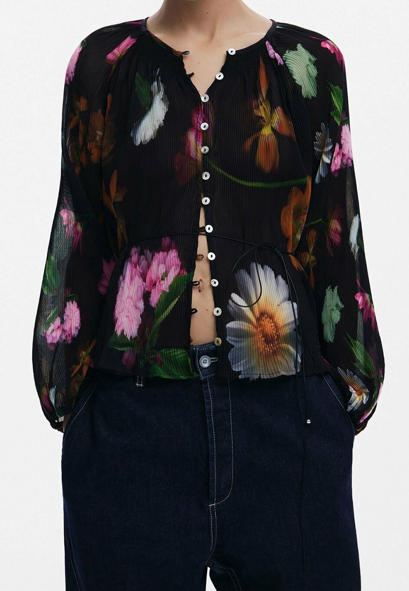 Blouse longue noire en tissu transparent avec imprimé floral multicolore, boutonnée devant, taille cintrée, portée avec un jean taille haute bleu foncé.