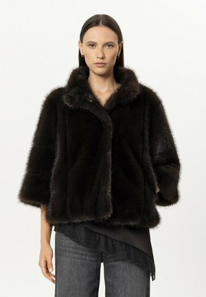 Woman with straight dark hair wearing a black fur jacket over a black top and gray jeans, standing against a plain white background.