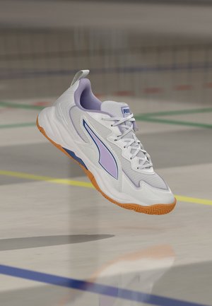 Λευκό και λεβαντίνι αθλητικό παπούτσι Puma με λαστιχένια σόλα να αιωρείται πάνω από πάτωμα γυμναστηρίου που έχει σημαδευτεί με πράσινες, κίτρινες και μπλε γραμμές.