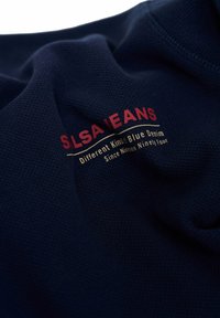 Tejido de mezclilla azul marino con un tejido texturizado. Presenta letras rojas y doradas que dicen "SLSA JEANS" y texto adicional debajo.