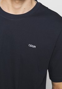 Nærbillede af en person iført en sort T-shirt med et lille, omvendt hvidt "HUGO" logo på venstre bryst.