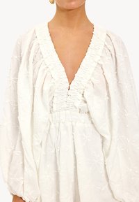 Witte jurk met diepe V-hals, gerimpelde halslijn en puffmouwen. Bevat bloemenbroderie en een elastische taille voor een ontspannen fit.