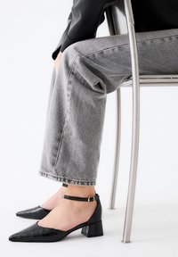 Zapatos de tacón bloque de charol negro con punta afilada y tira en el tobillo, con un patrón de cocodrilo texturizado. Combinados con denim gris.
