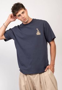 Marinblå bomullst-shirt med avslappnad passform, med en liten gyllene grafik på vänstra bröstområdet. Kortärmad och med rund halsringning.