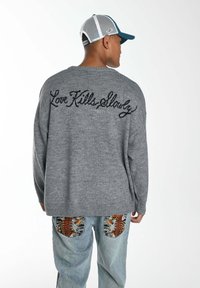 Grauer Strickpullover mit der Stickerei 'Love Kills Slowly' auf dem Rücken. Kombiniert mit hellblauen Jeans, die farbenfrohe gemusterte Taschen haben.