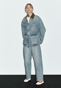 Denimjack met patchzakken en een lichtbruine boord van corduroy, gecombineerd met loszittende lichtblauwe jeans. De schoenen zijn witte flats.