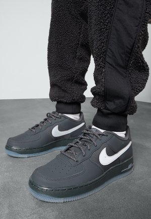 Harmaat nahkaiset Nike Air Force 1 -tennarit, joissa on valkoinen swoosh-logo, rei'itetty varvasosa ja läpinäkyvä kumipohja, yhdistetty mustiin teksturoiduihin collegehousuihin.