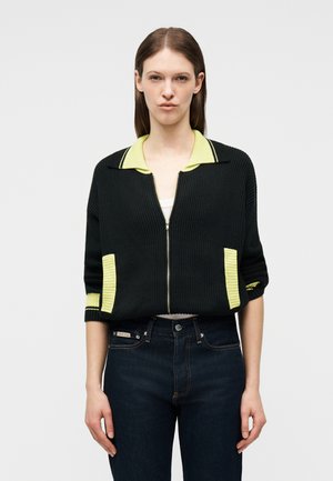 Mujer con cárdigan negro con cremallera, detalles en el cuello y bolsillo de color amarillo, combinada con jeans azul oscuro, posando frente a un fondo liso.