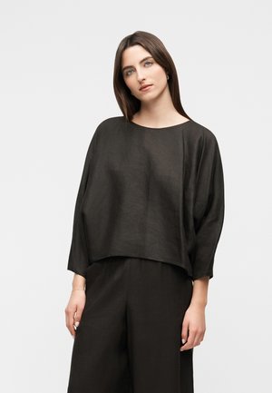 Mujer con cabello castaño lacio, vestida con un top negro de manga larga y pantalones anchos a juego, de pie contra un fondo claro y liso.