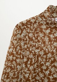 Blouse marron avec motif floral blanc, col montant à volants et plis verticaux sur l'épaule et la partie supérieure de la poitrine.