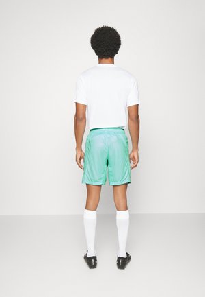 Man met krullend haar die met zijn rug naar de kijker staat, draagt een wit T-shirt, muntgroene korte broek, witte kniekousen en zwarte schoenen op een effen achtergrond.