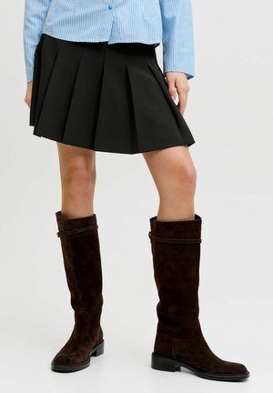 Femme portant une jupe plissée noire, une chemise à rayures bleues et des bottes en daim brun foncé montant jusqu'aux genoux, se tenant sur un fond clair uni.