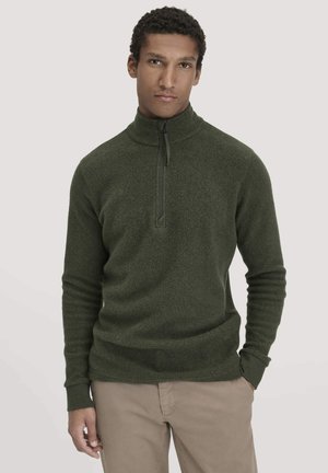 Man in olijfgroene fleece trui met kwartrits en beige broek, staande tegen een effen lichtgrijze achtergrond met handen in zijn zakken.