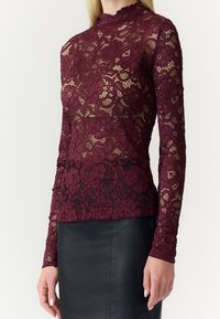 Femme portant un haut en dentelle florale bordeaux à manches longues sur une doublure nude, associé à une jupe noire ajustée.