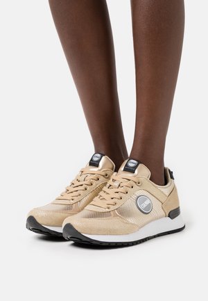 Chaussures de sport beiges avec une tige texturée, des accents en mesh, des détails noirs et un logo sur le côté. Elles possèdent des lacets et une semelle blanche.