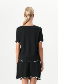 Žena s blond copom, oblečená v čiernom tričku s krátkym rukávom a sukni, obe s strieborným vlnitým lemom, zobrazená zozadu na bielom pozadí.