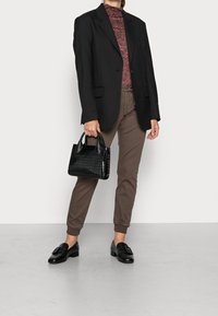 A person stands in a stylish black blazer and marbled red turtleneck, holding a small black handbag, with brown pants and shoes.