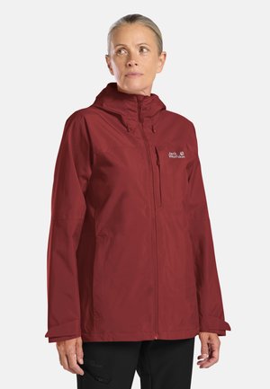 Frau trägt eine rote Jack Wolfskin Kapuzenjacke und schwarze Hose, schaut leicht zur Seite vor einem einfarbigen Hintergrund.