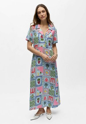 Vrouw draagt een kleurrijke, enkelhoge jurk met korte mouwen met geometrische en plantenpatronen, gecombineerd met witte, laaggehakte puntige schoenen.