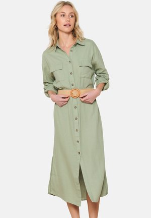 Vestito verde khaki con bottoni, dotato di due tasche sul petto, maniche lunghe ripiegate fino ai gomiti e una cintura larga intrecciata con una fibbia circolare.