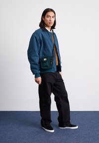 Chaqueta bomber azul, camiseta marrón, pantalones negros con bordado, zapatillas negras, bolsa verde con cremallera y logo. De pie sobre una alfombra azul.