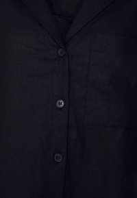 Chemise en lin noir avec un col cranté, une fermeture à boutons sur le devant et une poche plaquée sur le côté gauche. Texture lisse avec un léger éclat.