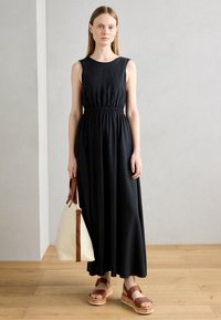 Black sleeveless maxi dress with gathered waist. Model holds a cream handbag with brown straps, wearing brown platform sandals with cork soles.