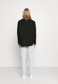 Camicia nera di lino con dettagli sui bottoni, indossata con pantaloni grigi chiari, abbinata a sneakers bianche, vista da dietro.