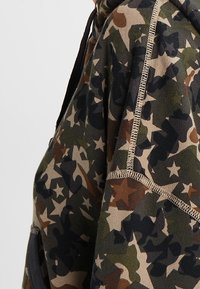 Sweatshirt à capuche dans un motif camouflage, featuring des tons verts, bruns et noirs avec des motifs d'étoiles. Tissu doux, coupe standard et couture visible.