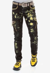 Czarne spodnie cargo z żółtymi wzorami tie-dye, złotymi ćwiekami, w zestawie z paskiem z ćwiekami oraz jaskrawymi żółto-czarnymi sneakersami.