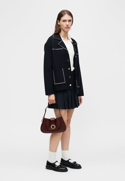 Γυναίκα που φοράει μαύρο τζάκετ με καρφιά, λευκό πουκάμισο, μαύρη πλισέ φούστα, λευκές κάλτσες, μαύρα μοκασίνια, κρατώντας καφέ τσάντα με χρυσό δαχτυλίδι.