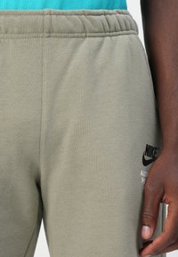 Grågrønne joggebukser med elastisk midjebånd; har et svart Nike-logo trykt på underbenet. Tyget er glatt og strukturert.