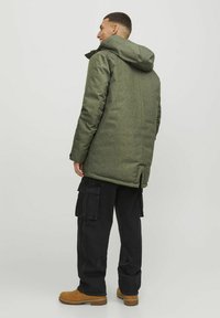 Zeleno, teksturirano dolgo plašč s kapuco, ki ima zadaj zračni vent. Nosil se je čez črne cargo hlače in rjave čevlje, kar prikazuje slojen videz.