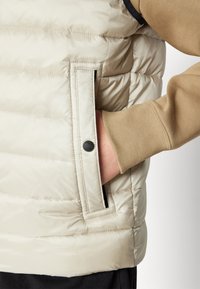 Beige gesteppte Weste mit einer Vorderseite, strukturiertem Stoff und einem schwarzen Knopfakzent. Brauner Ärmel eines Sweatshirts sichtbar.