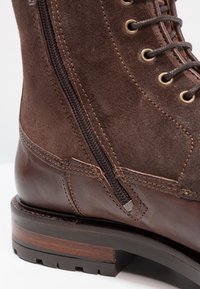 Brun mockasinankelstövel med dragkedja på sidan, läderdetaljer och en texturerad träklack. Har metallöglor och förstärkt sömnad.