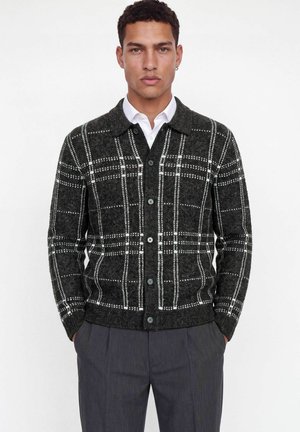 Homme portant un cardigan gris foncé boutonné avec un motif à carreaux blancs sur une chemise blanche et un pantalon gris foncé, debout les mains dans les poches.