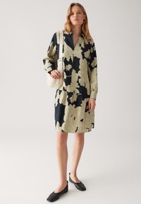 Frau in einem schwarz-beigen Blumenkleid, die eine weiße Umhängetasche trägt und schwarze Ballerinas an hat, steht vor einem schlichten hellen Hintergrund.