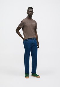 Bruine katoenen T-shirt met wit logo, blauwe spijkerbroek en groene sneakers met beige zolen. Model staat voor een neutrale achtergrond.