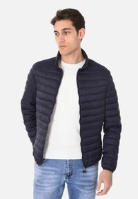 Giacca puffer navy con motivo a righe verticali, chiusura con zip e colletto alto, abbinata a un maglione bianco e jeans blu.