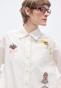 Bílá košile na knoflíky s třemi barevnými vyšívanými rostlinnými motivy: černý list, žlutý květ a detailní červený květinový vzor.