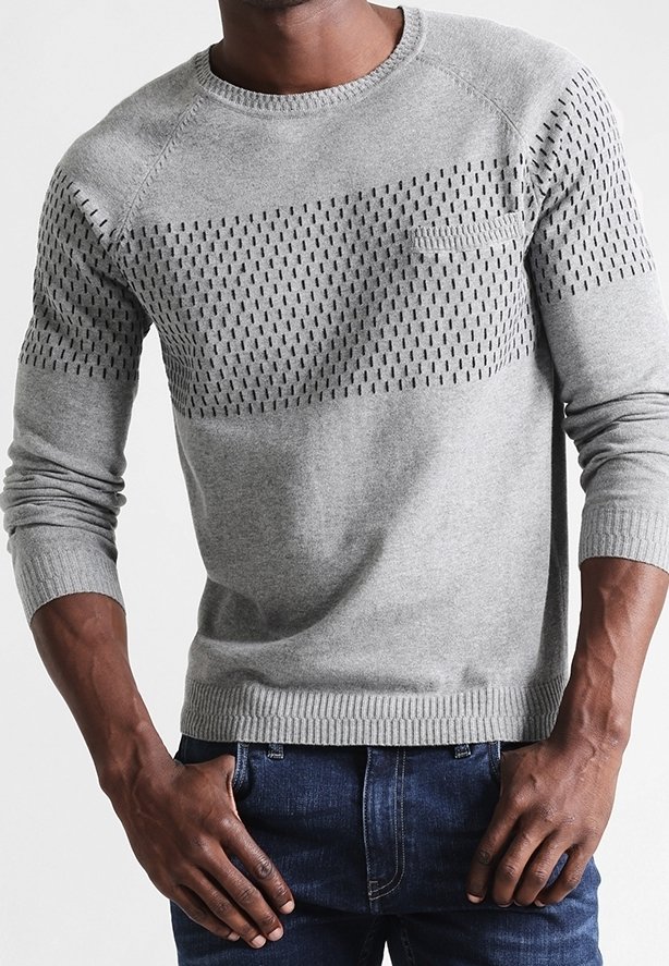 Homme portant un pull léger gris clair à col ras du cou avec de petites lignes noires verticales sur le haut de la poitrine et les manches, associé à un jean bleu foncé.