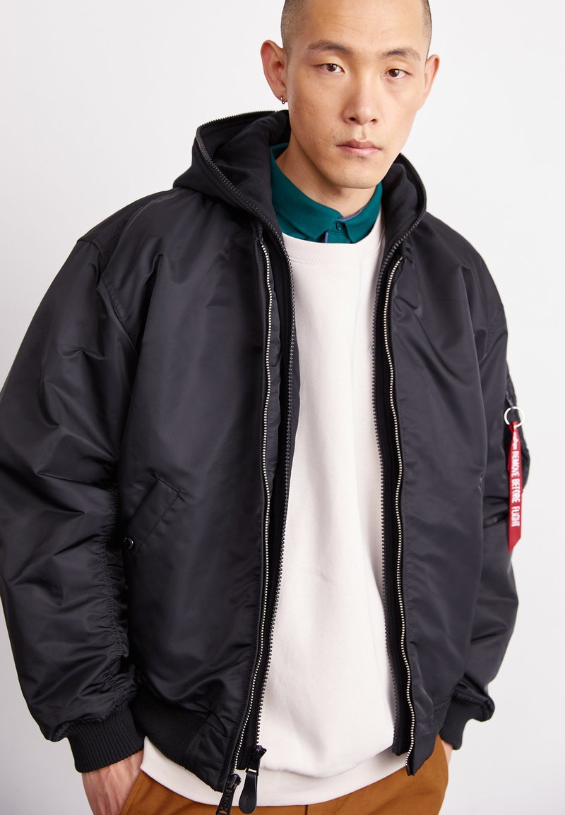 Veste bomber noire avec capuche, fermeture éclair, poignets côtelés et poches. Une étiquette rouge avec du texte est attachée à la fermeture éclair. Superposée à un pull crème.