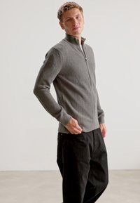Sudadera gris con cremallera, con un patrón texturizado, cuello alto y puños acanalados, combinada con pantalones negros sobre un fondo minimalista.