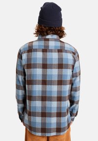 Camicia in flanella con motivo a quadri blu e marrone, dotata di colletto button-down. Indossata con un berretto lavorato a maglia di colore blu navy, caratterizzato da una vestibilità rilassata e un tessuto testurizzato.